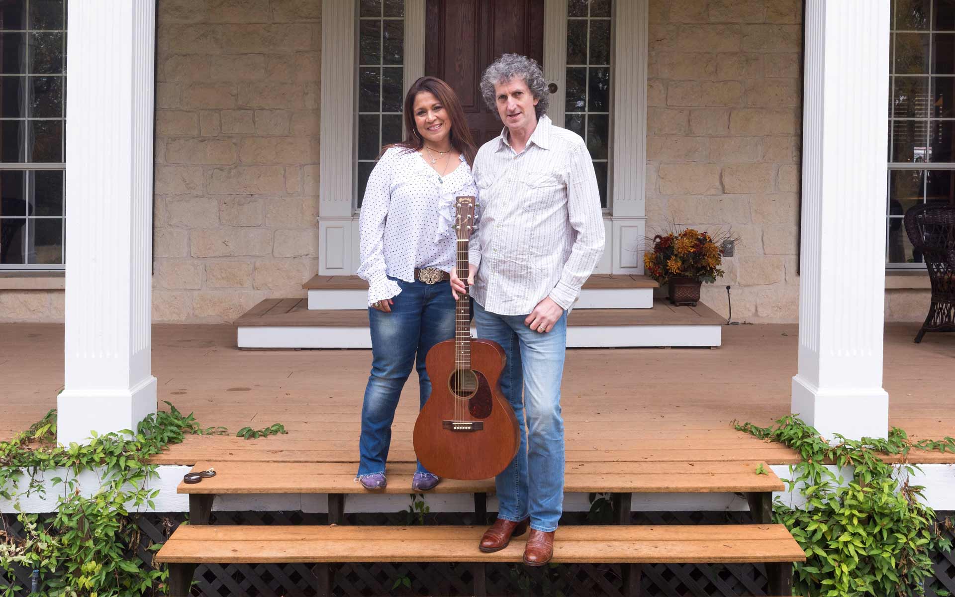 This is an image of Tommy & Saundra O'Sullivan standing together on the front porch of a home. They are smiling, and Tommy is holding an acoustic guitar, indicating their shared love of music.